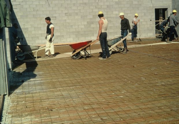 Men carrying concrete in wheelbarrows, pouring a reinforced concrete floor on corrugated metal on top of steel trusses.