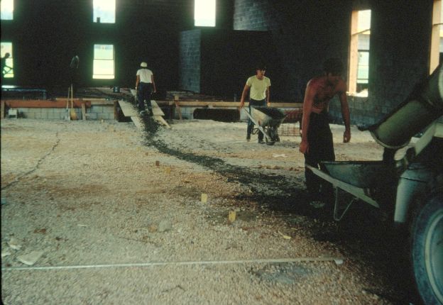 Men carrying concrete in wheelbarrows from a mixer truck to where a reinforced concrete floor is being poured.